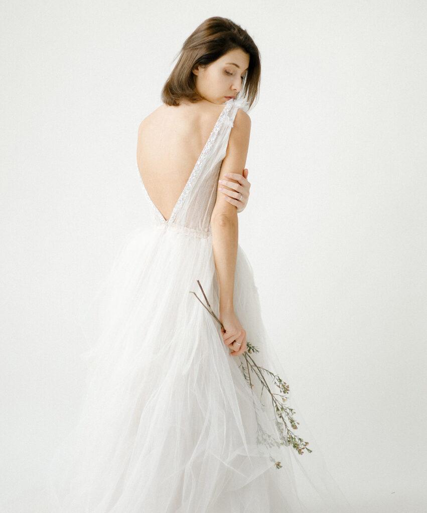Eine Frau trägt ein elegantes Brautkleid mit tiefem Rückenausschnitt und hält eine kleine Blumenstaude in der Hand. Ihr Blick ist nachdenklich.