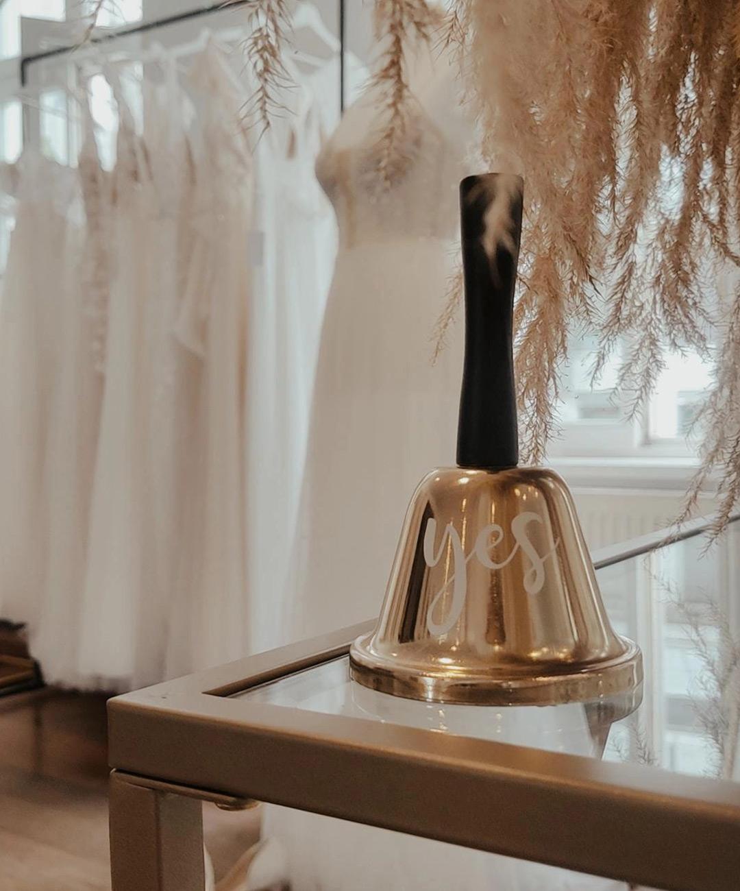 Eine goldene Klingel mit der Aufschrift 'yes' steht auf einem Tisch vor traumhaften Brautkleidern im Hintergrund.