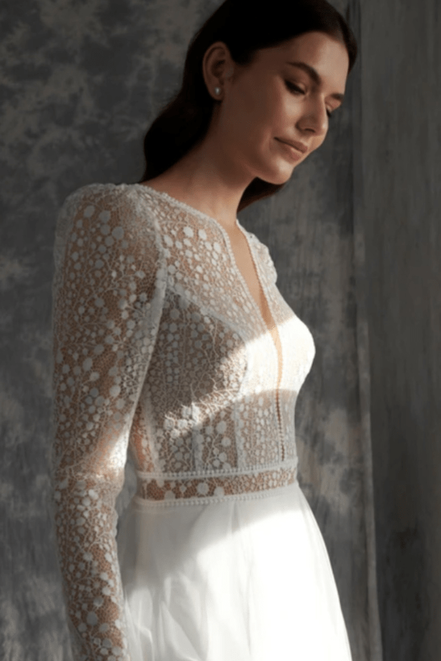 Eine Braut trägt ein elegantes Hochzeitskleid mit floraler Spitzenoberfläche und langem Arm. Die sanften Lichteffekte betonen die filigranen Details des Kleides.