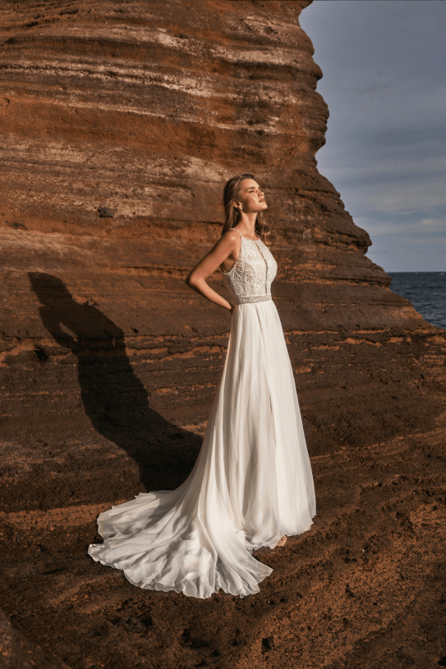 Eine elegante Braut in einem fließenden weißen Kleid steht vor einer Felswand am Meer. Ihr Kleid fällt sanft auf den Boden und die Sonne scheint auf sie.