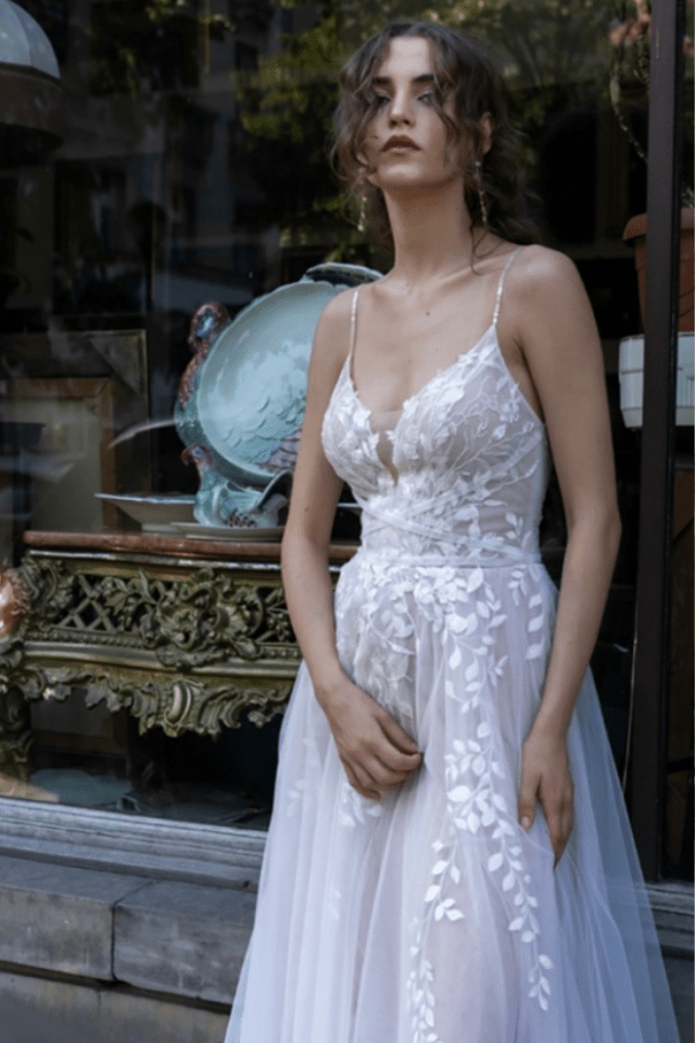 Eine Braut trägt ein elegantes, weißes Brautkleid mit floralen Spitzenapplikationen vor einem Schaufenster. Die feinen Details unterstreichen ihren persönlichen Stil.
