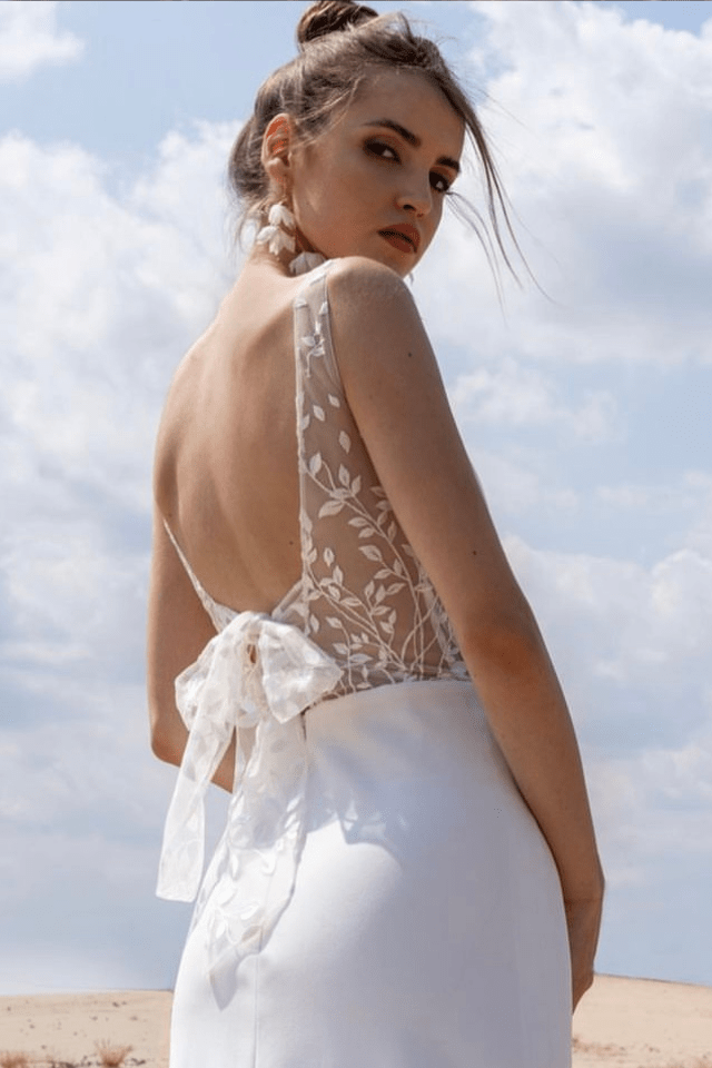 Eine junge Frau trägt ein elegantes Brautkleid mit Spitzenrücken und großen Ohrringen. Sie blickt über die Schulter, während der Himmel im Hintergrund klar ist.