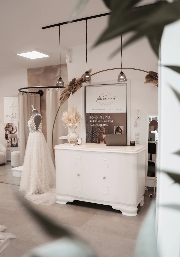 In einem Brautmodenstudio stehen elegante Brautkleider und eine weiße Kommode. Die Atmosphäre ist ruhig und einladend, ergänzt durch Accessoires wie Blumen.