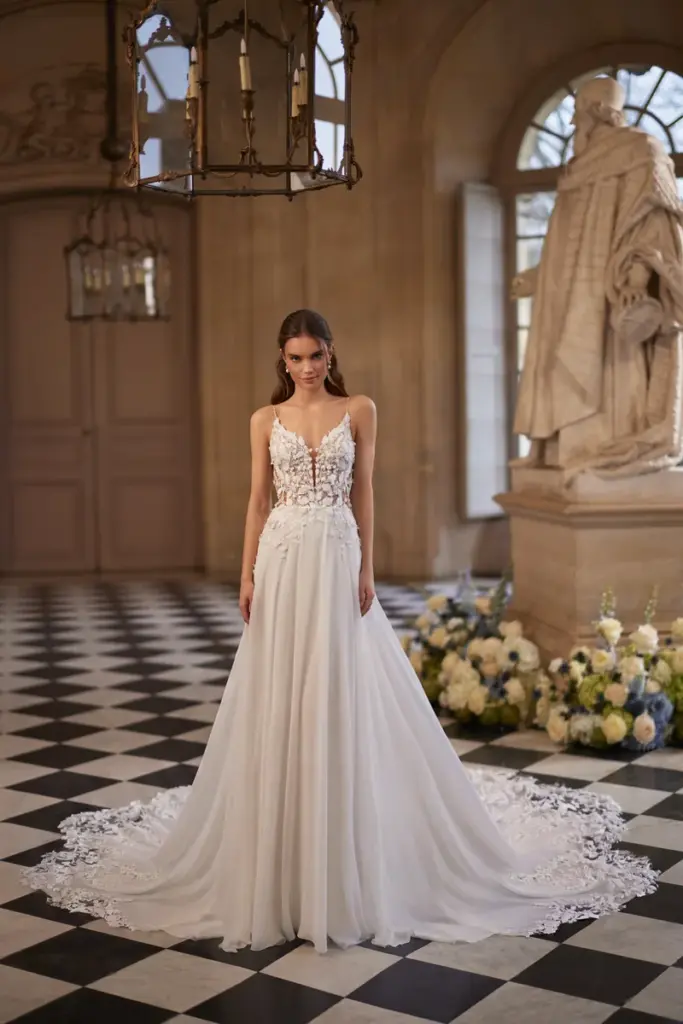 Eine Braut in einem Brautkleid mit schmalen Trägern, tiefem V-Ausschnitt und dreidimensionaler Blütenspitze am Oberteil; der weite Tüllrock endet in einer Schleppe mit Spitzensaum.