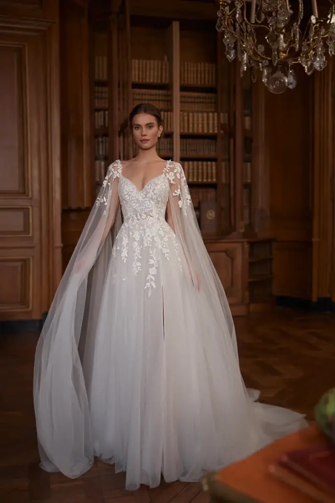 Ein Tüllkleid mit blattförmigen Spitzenapplikationen am Oberteil, hohem Beinschlitz und transparentem Cape über den Schultern.
