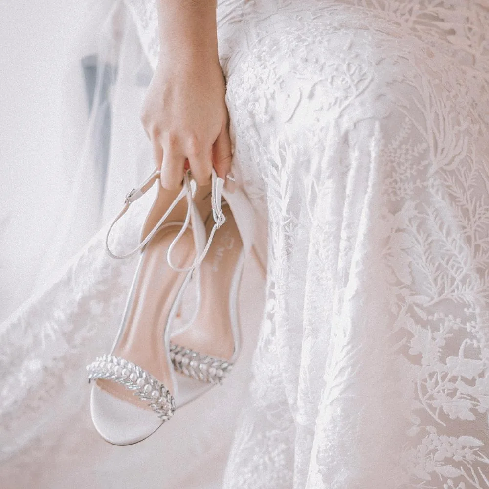 Eine Person hält elegante, weiße Brautschuhe mit Glitzerapplikationen in der Hand, während sie ein detailreiches, blütenweises Hochzeitskleid trägt.