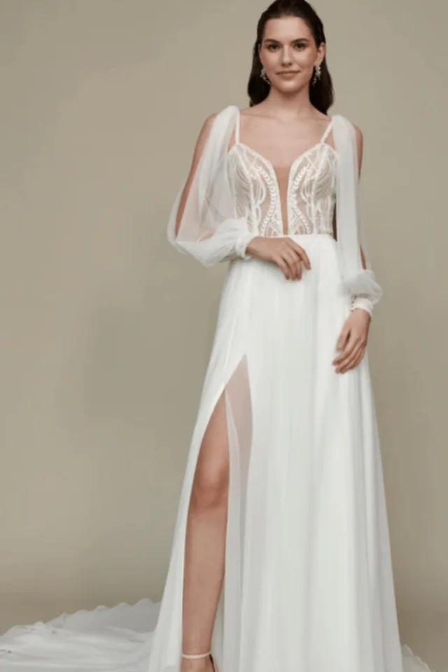 Eine Frau trägt ein elegantes Brautkleid mit transparenter Bluse und seitlichem Schlitz. Das Design verbindet modernes und klassisches Element.