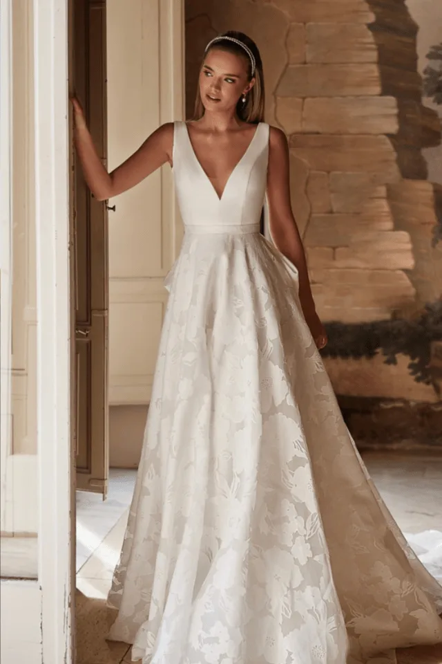 Eine Braut trägt ein elegantes, weißes Brautkleid mit tiefem V-Ausschnitt und floralem Muster. Sie steht vor einer Tür und lächelt.