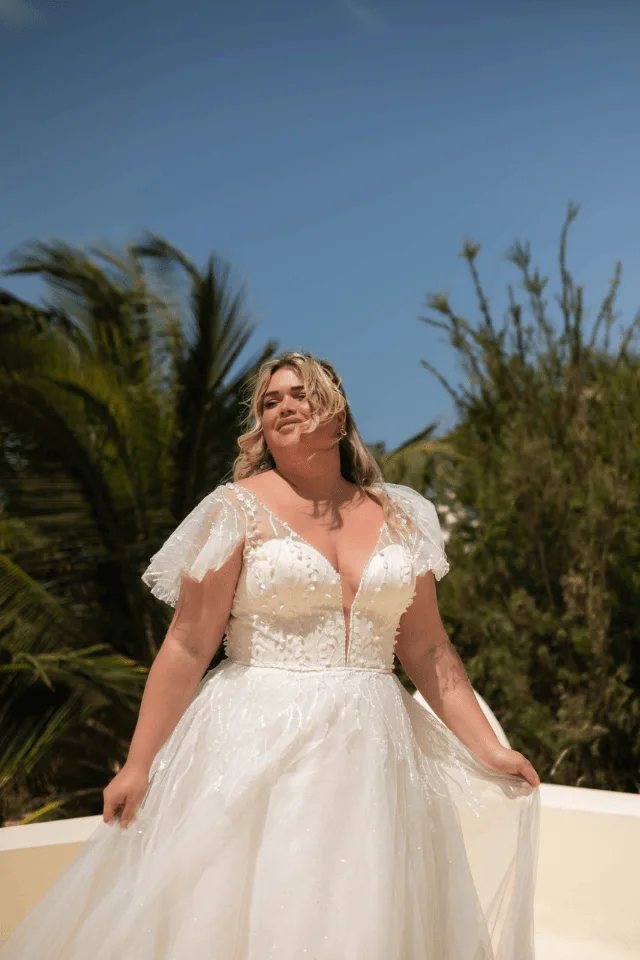 Eine Frau in einem eleganten, weißen Brautkleid mit Tüllärmel steht lächelnd vor einer grünen Landschaft; das Kleid hat feine Details und glitzert im Sonnenlicht.