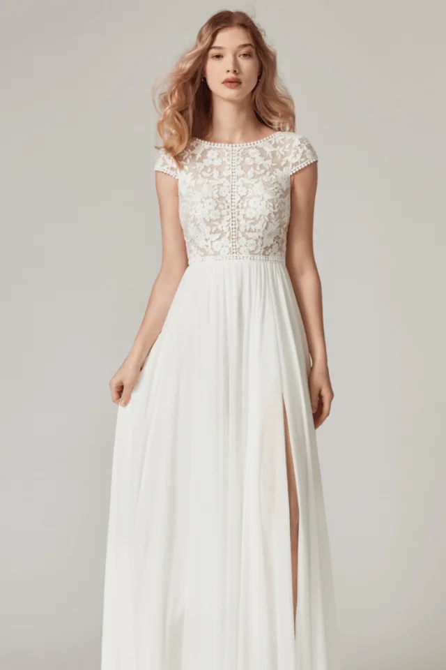 Eine Frau trägt ein elegantes, weißes Brautkleid mit floralem Spitzenoberteil und bequemer Rockpartie. Das Kleid hat einen Schlitz und betont die Figur.