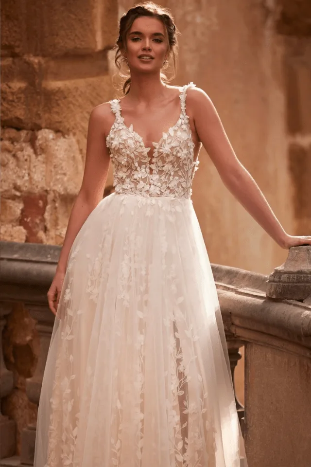 Eine Braut in einem eleganten, floralen Brautkleid steht an einer Treppe. Das Kleid hat transparente Details und zarte Spitzenapplikationen, die feminin wirken.