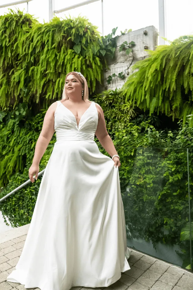 Eine Frau steht in einem eleganten, weißen Brautkleid vor einer grünen Pflanzenwand. Ihr Lächeln strahlt Freude und Selbstbewusstsein aus.