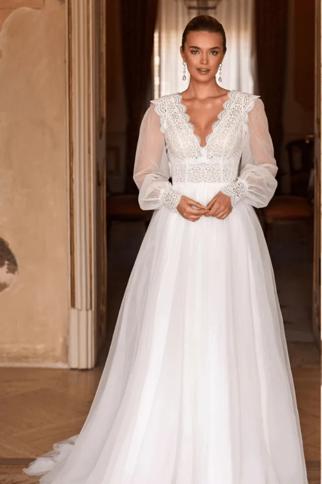 Eine Braut in einem eleganten, weißen Brautkleid mit Spitzendetails und langen, transparenten Ärmeln. Das Kleid hat eine romantische Silhouette, die die Persönlichkeit der Braut unterstreicht.