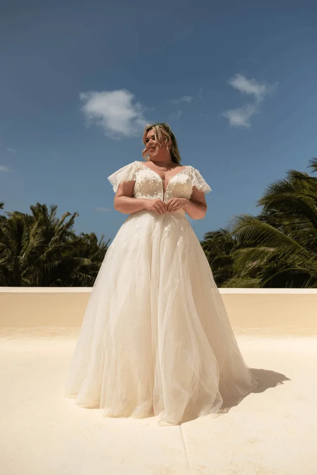 Eine Frau in einem eleganten, weißen Brautkleid mit tiefem Ausschnitt und schwingendem Rock steht auf einer Terrasse vor einem Strauch mit Palmen im Hintergrund.