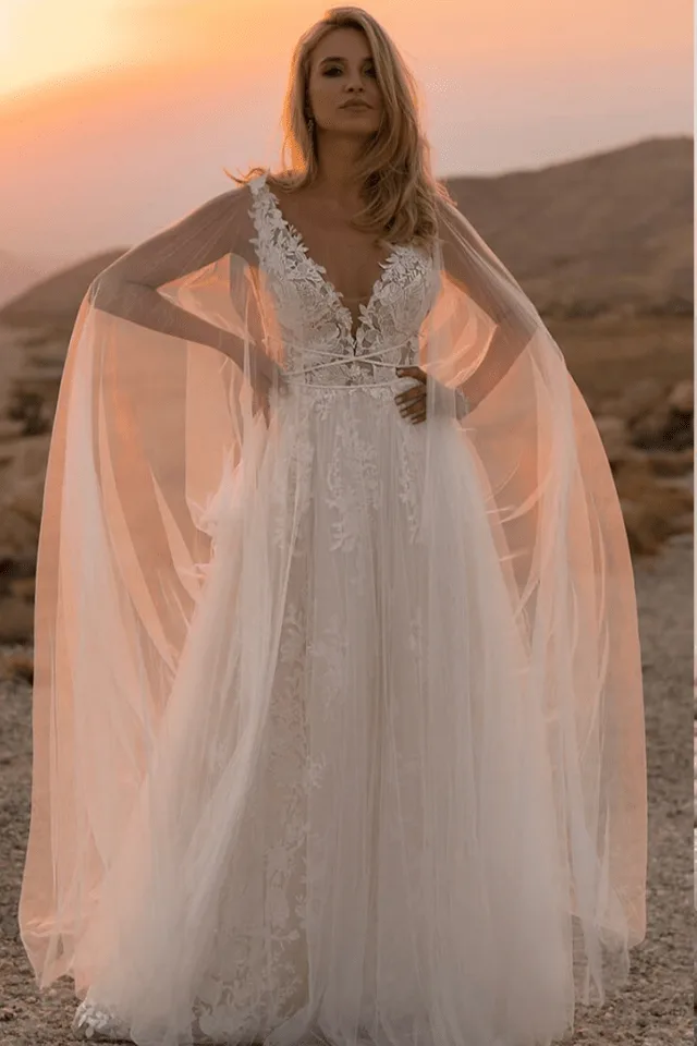 Eine Braut trägt ein elegantes, weißes Hochzeitskleid mit Spitzenakzenten vor einer dramatischen landschaftlichen Kulisse bei Sonnenuntergang.