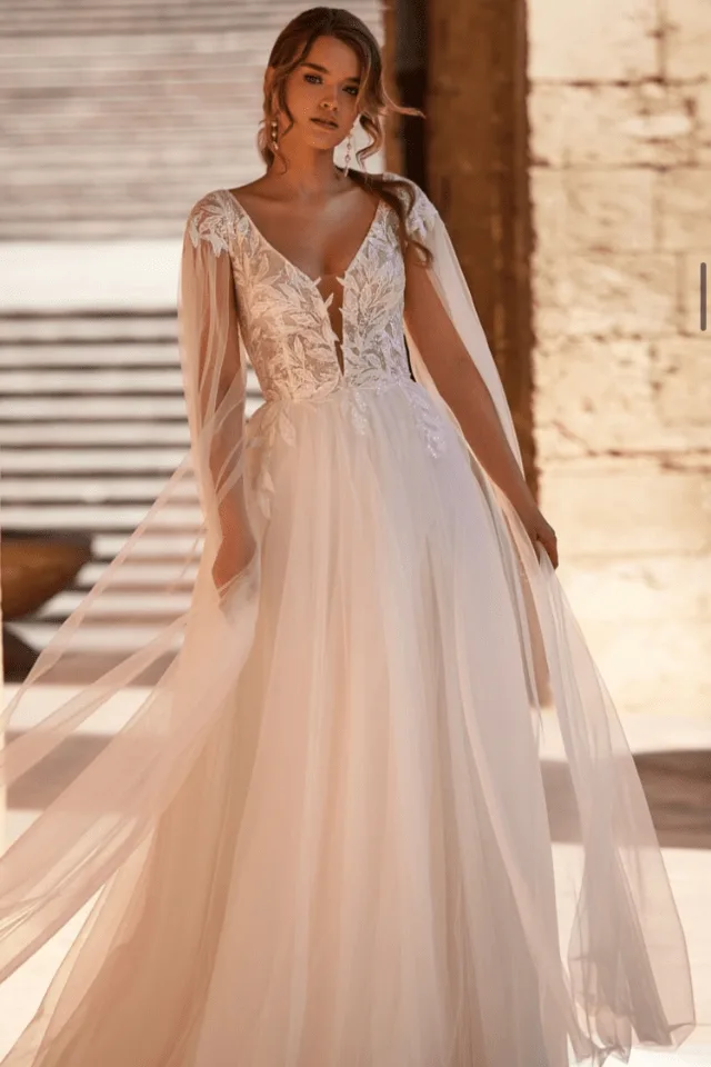 Eine Braut trägt ein elegantes, weißes Brautkleid mit transparenten Ärmeln und floralen Applikationen. Sie steht in einem hellen, rustikalen Raum.