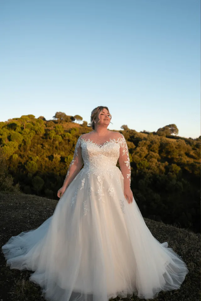 Eine Braut in einem eleganten, weißen Hochzeitskleid mit Spitze steht vor einer grünen Landschaft und lächelt. Ihre Pose strahlt Selbstbewusstsein und Freude aus.