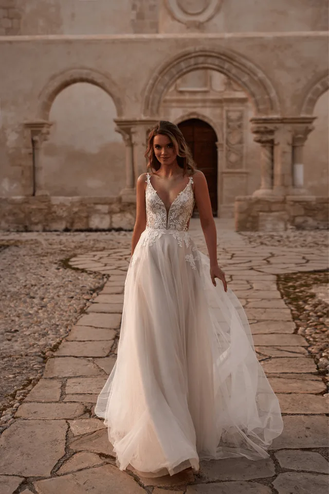 Eine Braut in einem eleganten, schlichten Kleid mit Spitzenoberteil geht auf einem gepflasterten Weg vor einem historischen Gebäude.