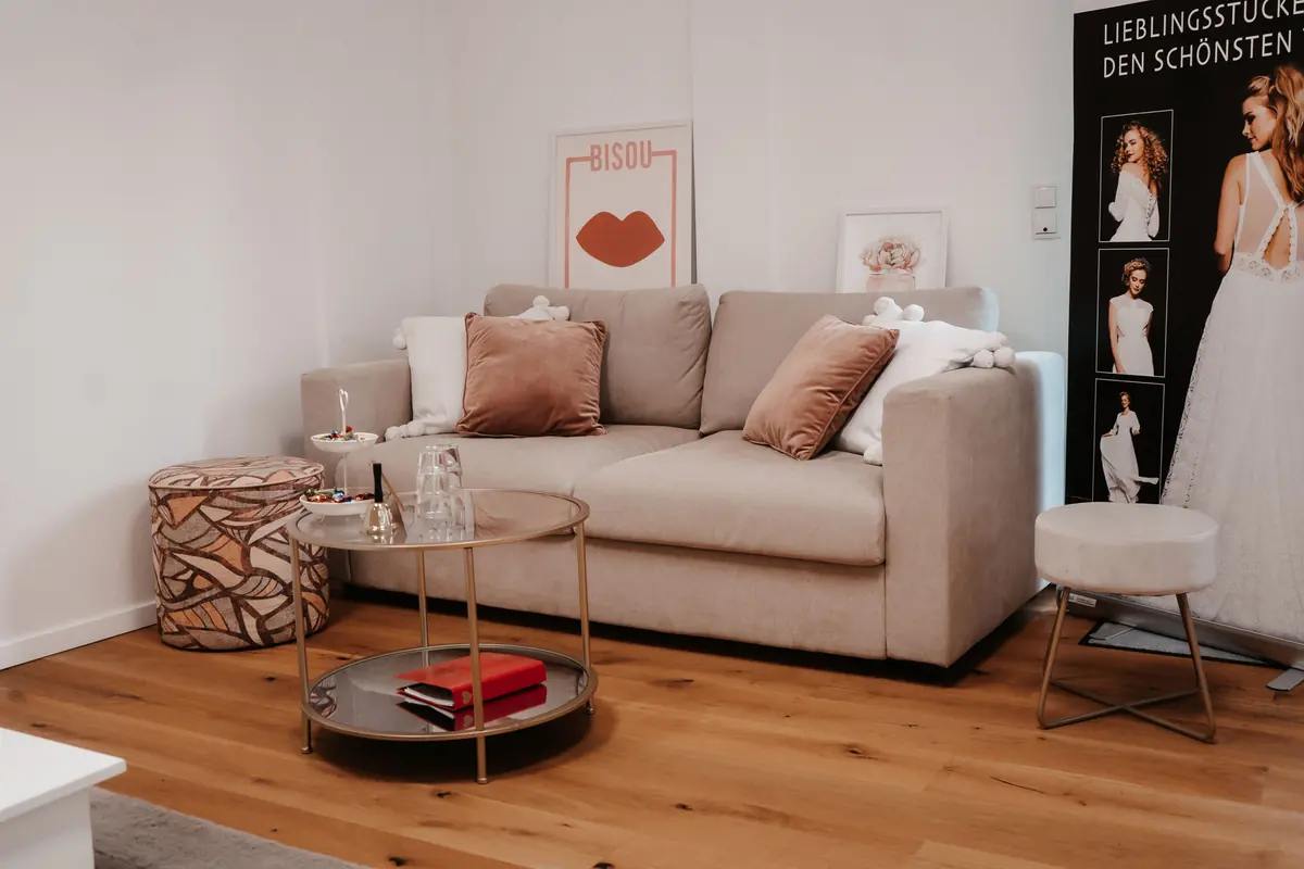 Sitzecke im Studio von Glücksmoment Brautmode Oldenburg mit einem hellen Sofa, Kissen und einem kleinen Beistelltisch — hier sitzt deine Begleitung während der Anprobe.